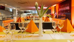 Ein Tisch in einer Uni-Mensa gedeckt mit Tischdecke, Weingläsern und orangenen Servietten 